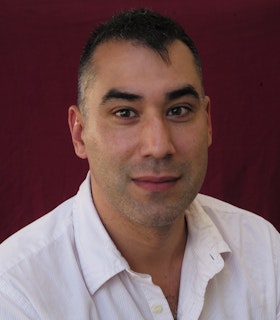 A portrait of Cedar Sigo, smiling, against a red background. He wears a white shirt and has short, dark hair. 