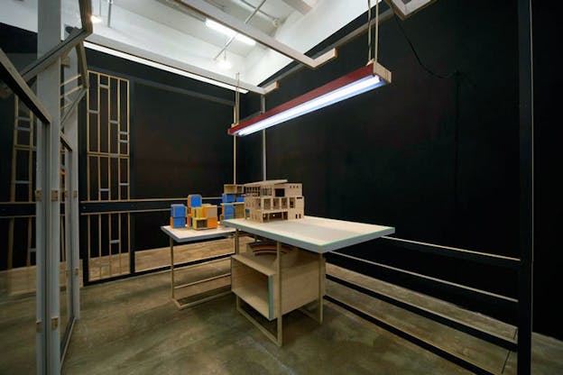 An installation image of a white L shaped table within a black walled room. On the left side of the table, numerous building blocks colored blue, yellow, and orange are stacked. On the right of the table there is a small wooden architectural model of a house. A red lamp hangs from the ceiling. 