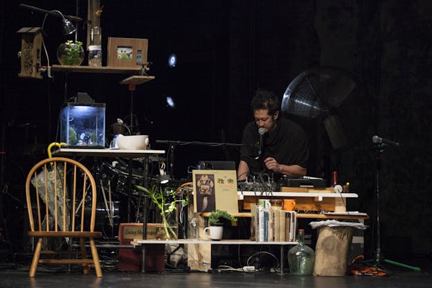 Tei Blow sits behind a coffee table covered in books and equipment next to shelving that holds small fishtanks, planters, and a birdhouse. He is in front of a black background, leaning over to speak into a microphone while his hands adjust controls on a sound board.