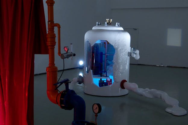 An installation image of a white cylindrical object in the center of a room. An abstract shape is cut out of the object, revealing a blue lit interior filled with purple pipes. The outside of the object is adorned with lightbulbs, an oval window pane, and an organically shaped, textured area made of the same white material. In the foreground, there is a red curtain and a red and blue pipe.  