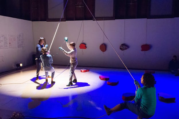 Four figures stand in a room with red flat shapes on the walls and floor, papers glued on a wall and ropes hanging from the ceiling. Three of them hold on to a rope while a fourth holds on its opposite end.