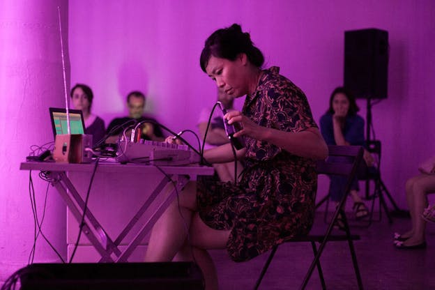 A figure wearing a patterned dress sits in front of a desk holding a small electronic box connected to the electronic in front of them, in a purple lit room. An audience watches them from the sidelines.