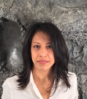 Athena LaTocha stands in front of a stony gray background and looks directly at the camera. She is dressed in a white button up blouse. 