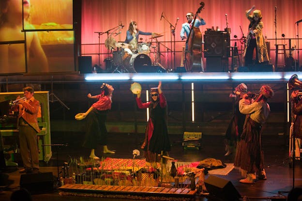 Performers, some dressed in traditional Sufi garb, others in suits and ties play various instruments on a double-leveled stage with a screen projection in the upper left corner of a child. 