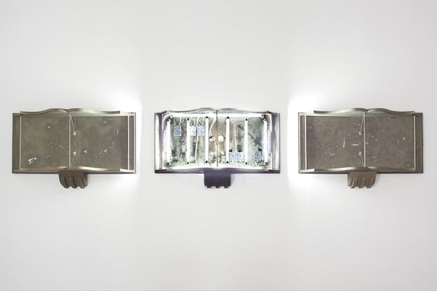 Three aligned frames on a wall in the outline of books. Two on either side sit empty with a gray-brown background, while the one in the middle has white long lights and cables.