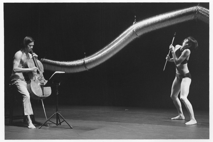 A black and white photograph of two performers in front of a long and curved aluminum duct hung from above. The performer on the left wearing a pair of pants plucks the strings of a cello while looking down at the music stand. The performer on the right wearing a bralette and a brief underwear holds a cello bow while tilting their head to the right and slightly bending their knees. The stage has a light color floor and a dark backdrop.