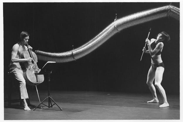 A black and white photograph of two performers in front of a long and curved aluminum duct hung from above. The performer on the left wearing a pair of pants plucks the strings of a cello while looking down at the music stand. The performer on the right wearing a bralette and a brief underwear holds a cello bow while tilting their head to the right and slightly bending their knees. The stage has a light color floor and a dark backdrop.