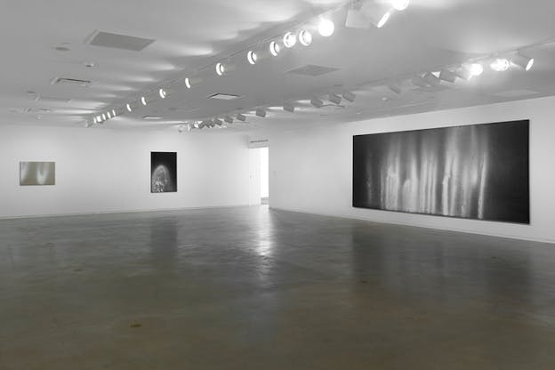 A gallery space of white walls holds on its right a black canvas with white grainy ombre lines. In the left sit two smaller beige and black canvases with the same white grainy effect.
