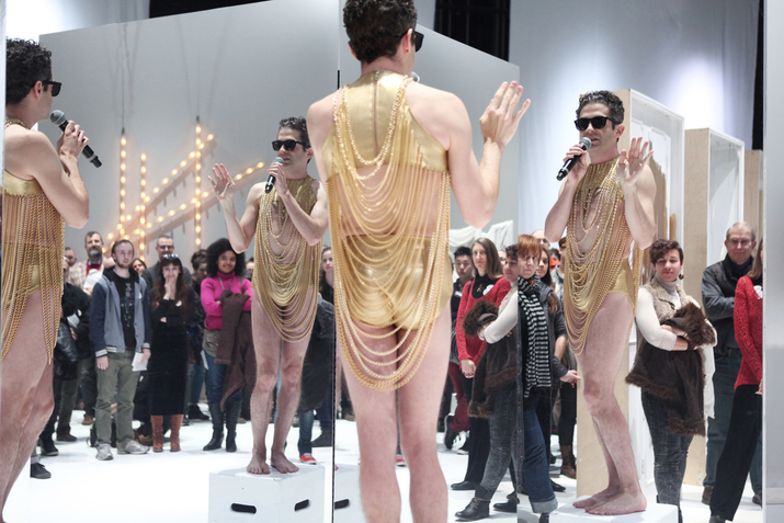 A person standing on white step stool, dressed in black sunglasses, metallic gold underwear and crop top, with pearls starting from the neckline and falling down in a 'U' manner talks in a microphone. Their reflection is shown four times on full body mirrors, as a crowd watches. 