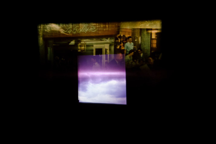 Rectangle projecting imagery of clouds blending into a transparent violet rectangle superimposed on a dim green and yellow hued rectangular image of people gathered at an indoor space marked by a sign reading 