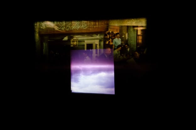 Rectangle projecting imagery of clouds blending into a transparent violet rectangle superimposed on a dim green and yellow hued rectangular image of people gathered at an indoor space marked by a sign reading 