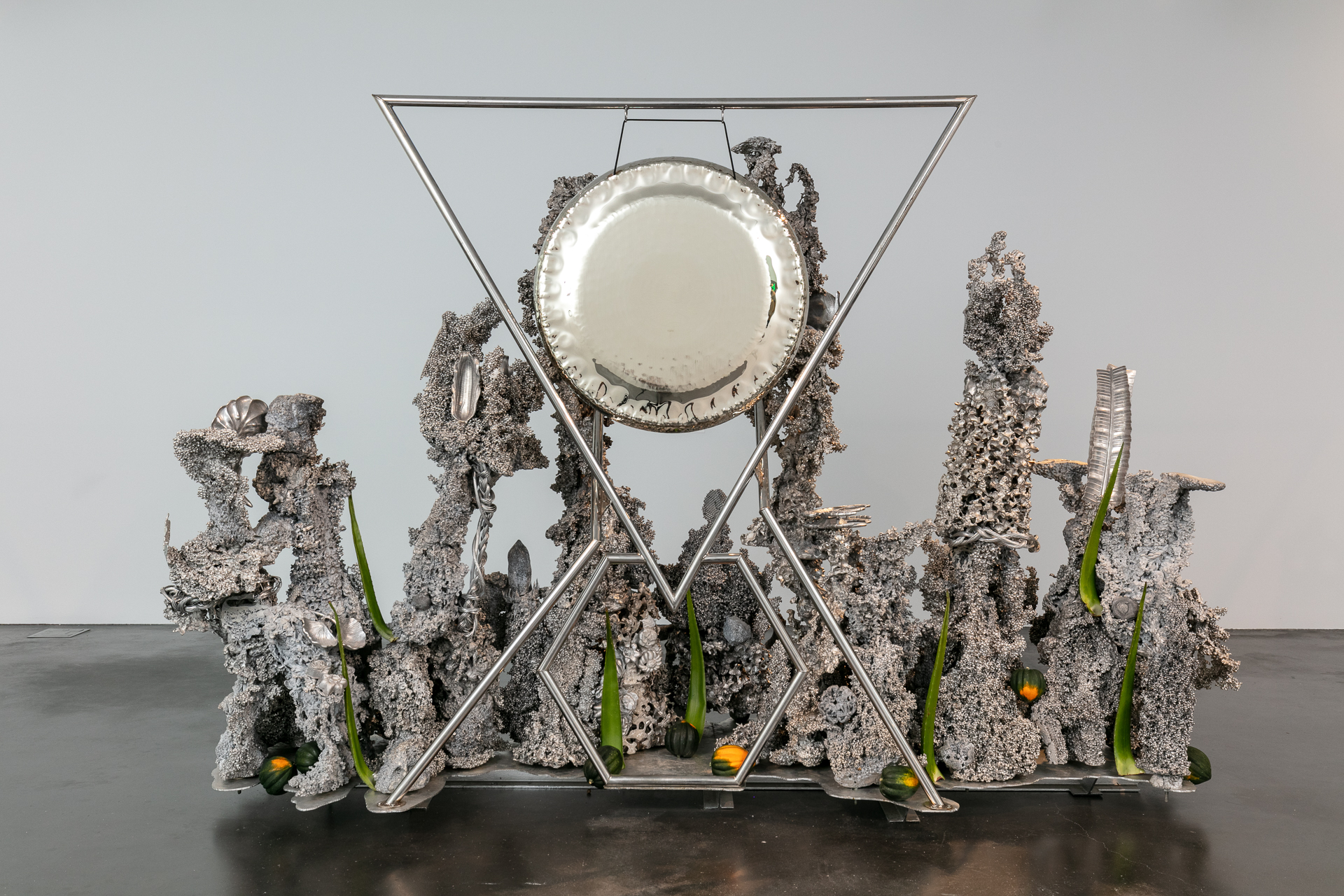 A wide silver metallic sculpture with a large silver plate hanging suspended in a triangular metal frame, braced point down above a smaller hexagonal frame. Surrounding the metal frame are various textured tendrils of welded silver material that twist upward. The tendrils have a rough, granular texture and are ornamented with braided wires and silver metal moulded in the form of various objects, including gingko leaves, palm fronds, an ear of corn, seashells, and flowers. Stalks of aloe vera are affixed among the metal tendrils, pointing upwards. Small acorn squashes are spread around the base of the sculpture.