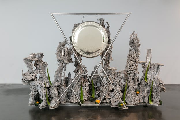 A wide silver metallic sculpture with a large silver plate hanging suspended in a triangular metal frame, braced point down above a smaller hexagonal frame. Surrounding the metal frame are various textured tendrils of welded silver material that twist upward. The tendrils have a rough, granular texture and are ornamented with braided wires and silver metal moulded in the form of various objects, including gingko leaves, palm fronds, an ear of corn, seashells, and flowers. Stalks of aloe vera are affixed among the metal tendrils, pointing upwards. Small acorn squashes are spread around the base of the sculpture.