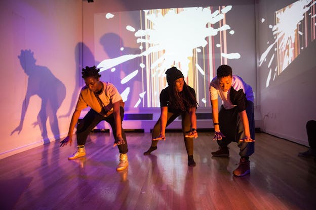 Three performers lean over, extending their arms so that they nearly touch the wooden floor. Behind them, a projection against the wall shows a firework graphic against a series of multi-colored stripes. The room is lit with a light purple color, casting shadows of the performers onto the left wall. 