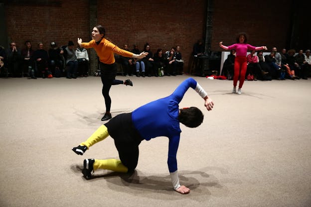 Three performers on a beige stage in different poses stand apart from one another. A performer clad in pink furthest away from the viewer holds their hands wide open, as another closer one dressed in orange moves forward with wide open arms and a raised leg. In the front closest to the viewer the third performer with their back turned and dressed in a blue shirt, black pants and yellow socks, supports themselves sideways with one arm and one leg.