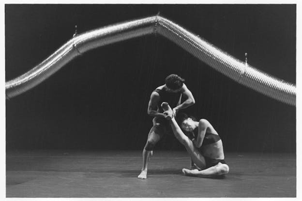 A black and white photograph of two performers in front of a long and curved aluminum duct hung from above. The performer on the right wearing a bralette and a brief underwear sits on the floor while raising their right leg straight up. They lean their body to their right with a supporting right arm. The performer on the left wearing rompers holds the other performer's raised leg while looking at them in a half squat position. The stage has a light color floor and a dark backdrop.