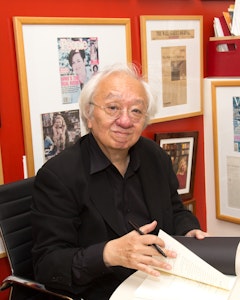 Portrait of a sitting John Yau with white ear-length hair, dressed in thin framed circular glasses and a black suit with a matching button-up underneath. He holds a folding paper and a pen in his hands on a desk where two wine glasses are situated. Behind him a red wall with frames of different images and articles.