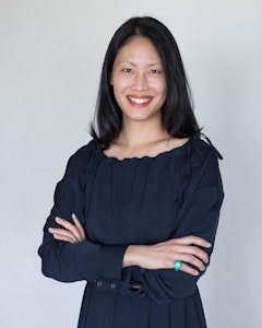 Portrait of a smiling Emily Wei Rales with long straight black hair and a dark blue long sleeve dress. The arms are folded in front and they fashion a bright blue ring on the pinky of the left hand.