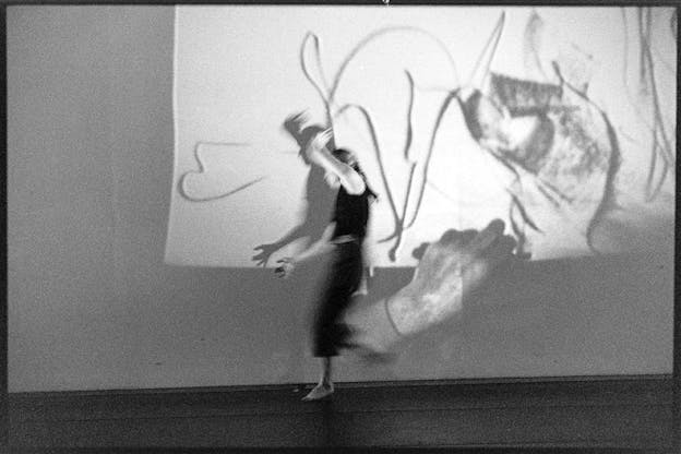 A performer dances in front of a projection of a hand drawing lines on paper. They wears a dark outfit and rest on one foot. One of their arms extends in front of them and the other is above them. 