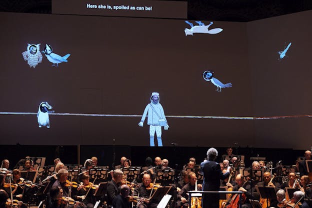 An orchestra performs as behind them on stage a projection of an astronaut surrounded by various animals and on top of them the phrase 