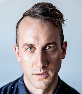 A cold color scheme muted portrait of Andrew Schneider dressed in a blue and white polka dot button up, with green eyes and a swept to the side hairstyle.