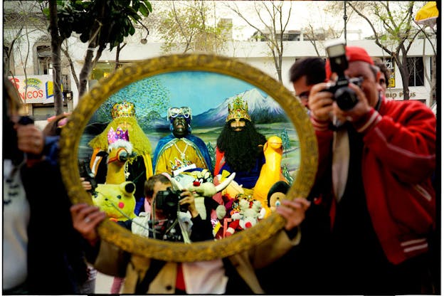 In the middle of a city street, there is a cluster of people. The one of the right wears a red jacket and holds up a camera to their eye. The person on the left covers their face with an oval gold framed image of people in play royalty costumes in front of mountains and surrounded by toys and a person taking a photograph. 
