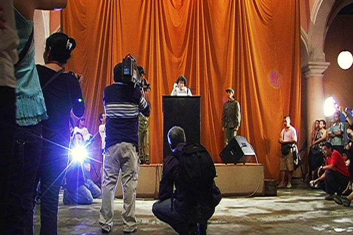 A figure behind a stand on a small stage with red drapes talks in a microphone while photographers and videographers stand opposite them with their back towards the viewer.