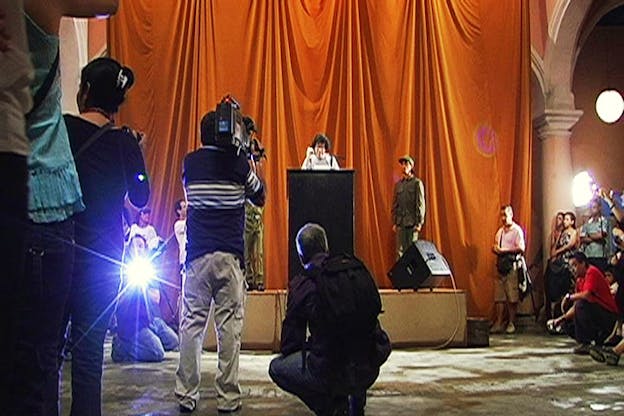 A figure behind a stand on a small stage with red drapes talks in a microphone while photographers and videographers stand opposite them with their back towards the viewer.
