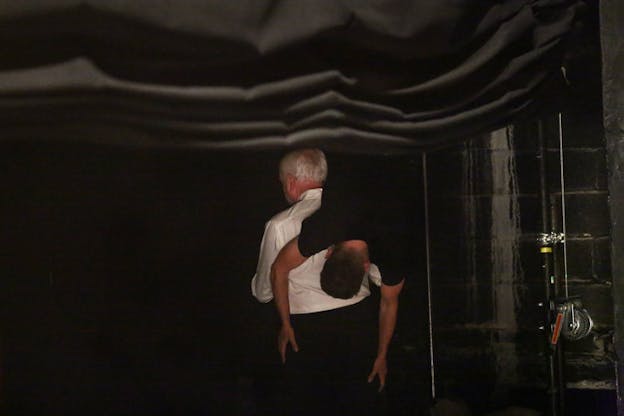 A person dressed in dark pants and a white top holds thrown over their shoulder a figure dressed in complete black. Both figures have their backs turned to the viewer.  