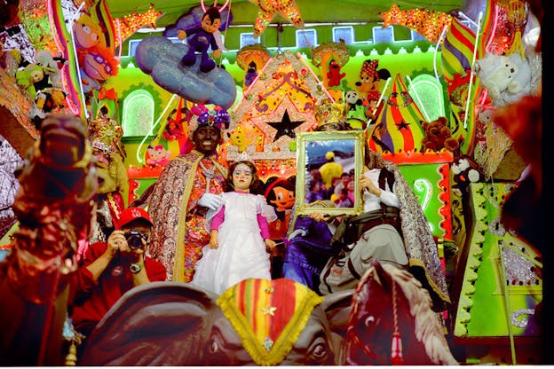 There is a neon green walled crowded space filled with toys and stuffed animals. The windows in the green wall are rounded-rectangle shaped windows and sparkly green trim. In the center of the green wall, there is a golden triangle that is covered in sparkly decorations. Under this triangle, a person wearing a plush joker hat and an elaborate cape holds the shoulders of a child beside them. The child wears a white fancy dress over fushia long sleeves, a tiara, and face paint. To the right of the child, a person wearing a white shirt holds a gold framed image. The image within the frame is brightly colored but blurred. 