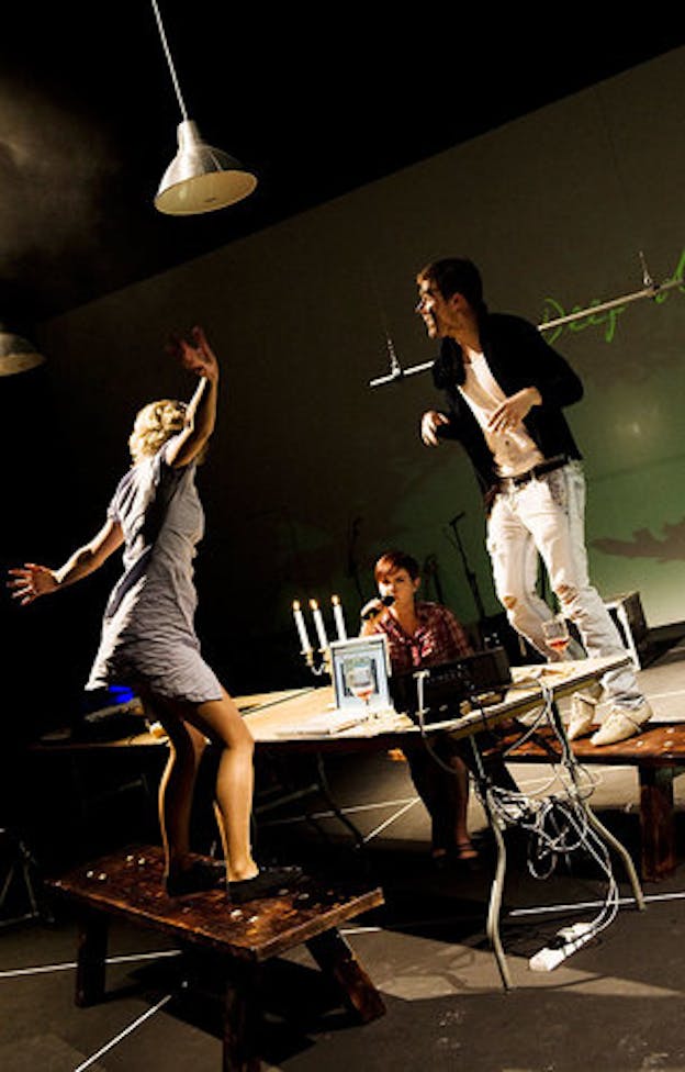 Three performers play instruments in a darkened space illuminated only by a projection which is composed of an abstract colored in background and white text. 