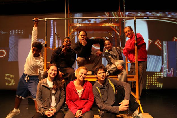 Several people sit around some scaffolding wearing casual clothes. They all look at the camera and smile. In the background, there is a large screen projection an abstract image. 