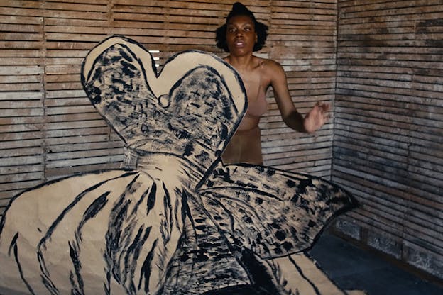 A performer in a wooden shed dressed in nude underwear, holds a cut out of a dress, its silhouette painted over with black swipes.