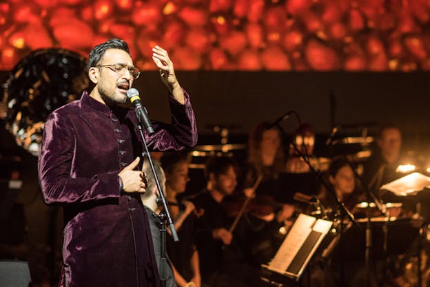 A performer dressed in purple velvet and glasses exclaims in a microphone. A blurred orchestra sits behind him.