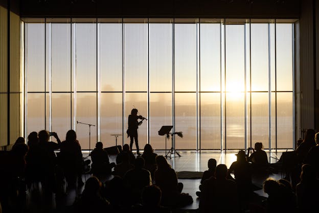 A photograph of Laura Ortman performing with her violin against a wall of windows that show a city skyline at sunset. She is backlit and so are the audience members sitting on the floor in front of her. 