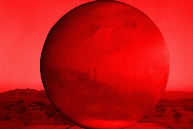 Red-hued image of a large planet-shaped orb set against a red desert with mountains in the far distance and a cloudless red sky. 