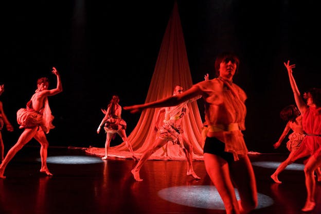 Performers on stage are illuminated by a red light. Behind them on the stage hangs a drape forming an upside down flower.