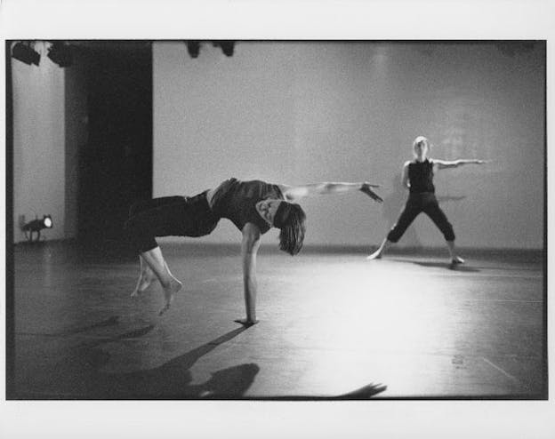 A performer in the foreground bends their body in a table top position over the stage. In the background, a different performer lunges and reaches their left arm outwards all while looking directly towards the camera.