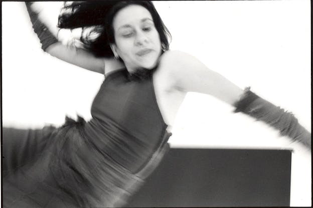 A black and white image of a dancer flinging themselves backwards. Their eyes are closed and their head is tilted downwards. Their right arm is flung upwards and their left arm reaches downwards. They have dark shoulder length hair and wear a dark halter neck and dark pants.  
