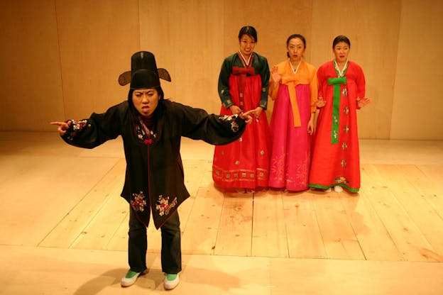 Three people dressed in traditional Korean wear stand in the background with exaggerated upset expressions. In the foreground, another person dressed in a black traditional Korean costume points their fingers outward while frowning.