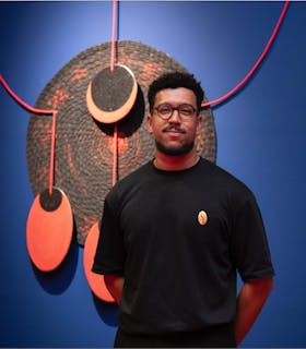 A portrait of Yhuri Cruz wearing a black t-shirt and standing with his arms crossed behind his back. He stands in front of a vibrant blue wall, upon which a layered, geometric wall sculpture comprised of circle and oval forms hangs. The portrait is framed by a soft mauve square. At the top it reads