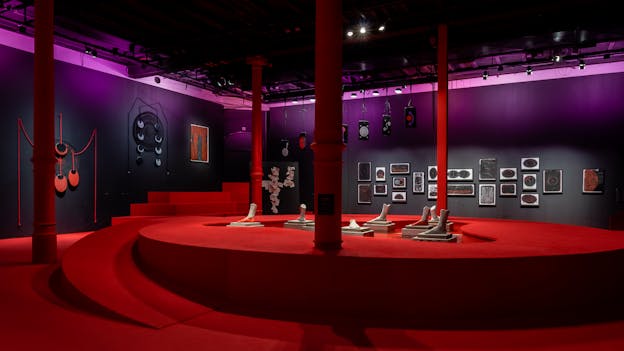 A gallery space containing an installation of works by Yhuri Cruz. The walls are painted black and are illuminated by dramatic purple lighting. There are several artworks throughout the space, including some framed drawings of abstract, otherworldly forms rendered in muted colors. The entire floor is covered in crimson red carpeting and in the center of the space there is a ramp that leads up to a circular stage. In the middle of the stage, several sculptures of individual feet reach rest on pedestals that emerge from the floor. Throughout the space are several large red support columns extending upwards to the ceiling.