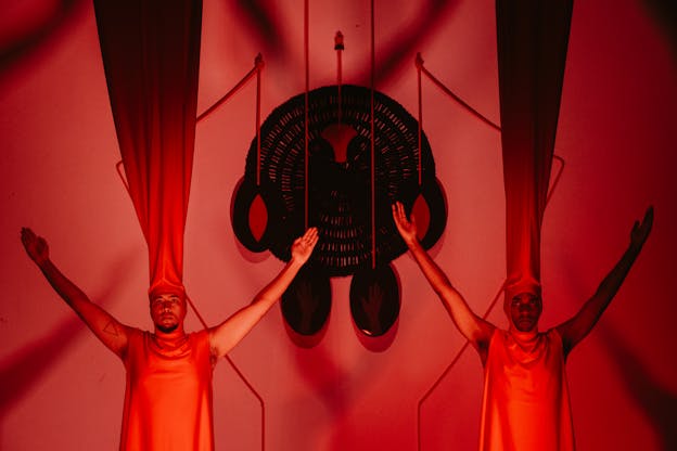 Two performers stand in the foreground facing the camera with their arms outstretched in the air. Both are clad in crimson red tunics and matching red headdresses with the fabric extending outwards from their heads towards the ceiling. The lighting in the scene is dramatic, with performers illuminated and the tops of their headdresses are covered in heavy shadow. The background is a deep red and suspended between them is a large geometric sculpture. The shadows from the performers’ positions are visible on the back wall.
