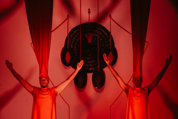 Two performers stand in the foreground facing the camera with their arms outstretched in the air. Both are clad in crimson red tunics and matching red headdresses with the fabric extending outwards from their heads towards the ceiling. The lighting in the scene is dramatic, with performers illuminated and the tops of their headdresses are covered in heavy shadow. The background is a deep red and suspended between them is a large geometric sculpture. The shadows from the performers’ positions are visible on the back wall.