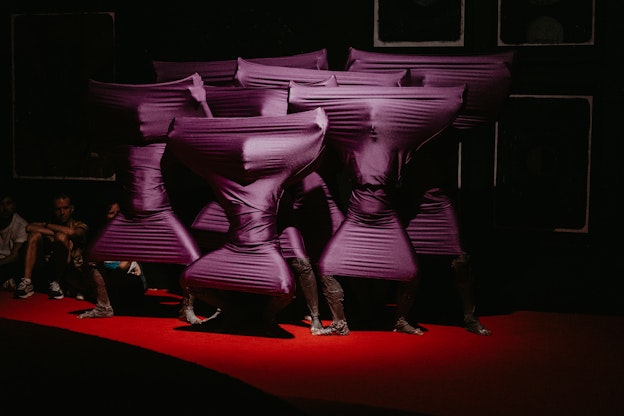 A group of performers are dressed in stretchy purple fabric garments that are wrapped around their bodies. They are each standing positioned in different poses with their arms and legs outstretched, resulting in geometric shapes emerging from the fabric. They stand atop a red stage and there are a couple figures seated on the floor directly behind them. The space is dimly lit and the performers are illuminated by a soft spotlight, conveying a theatrical scene.