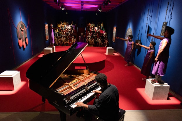 A performer dressed in a black suit sits in the foreground playing a grand piano with their back turned to the camera. Four additional performers are positioned across the room, with two standing atop white plinths and two on the floor. They are all dressed in matching sleeveless purple garments and have one arm raised towards the center of the room mid performance. The walls are painted in deep blue and there are abstract artworks hanging on the walls. Red carpeting covers the floor and a round, red platform is visible in the center of the image. At the back of the room there are two groups of spectators seated, watching the performance.