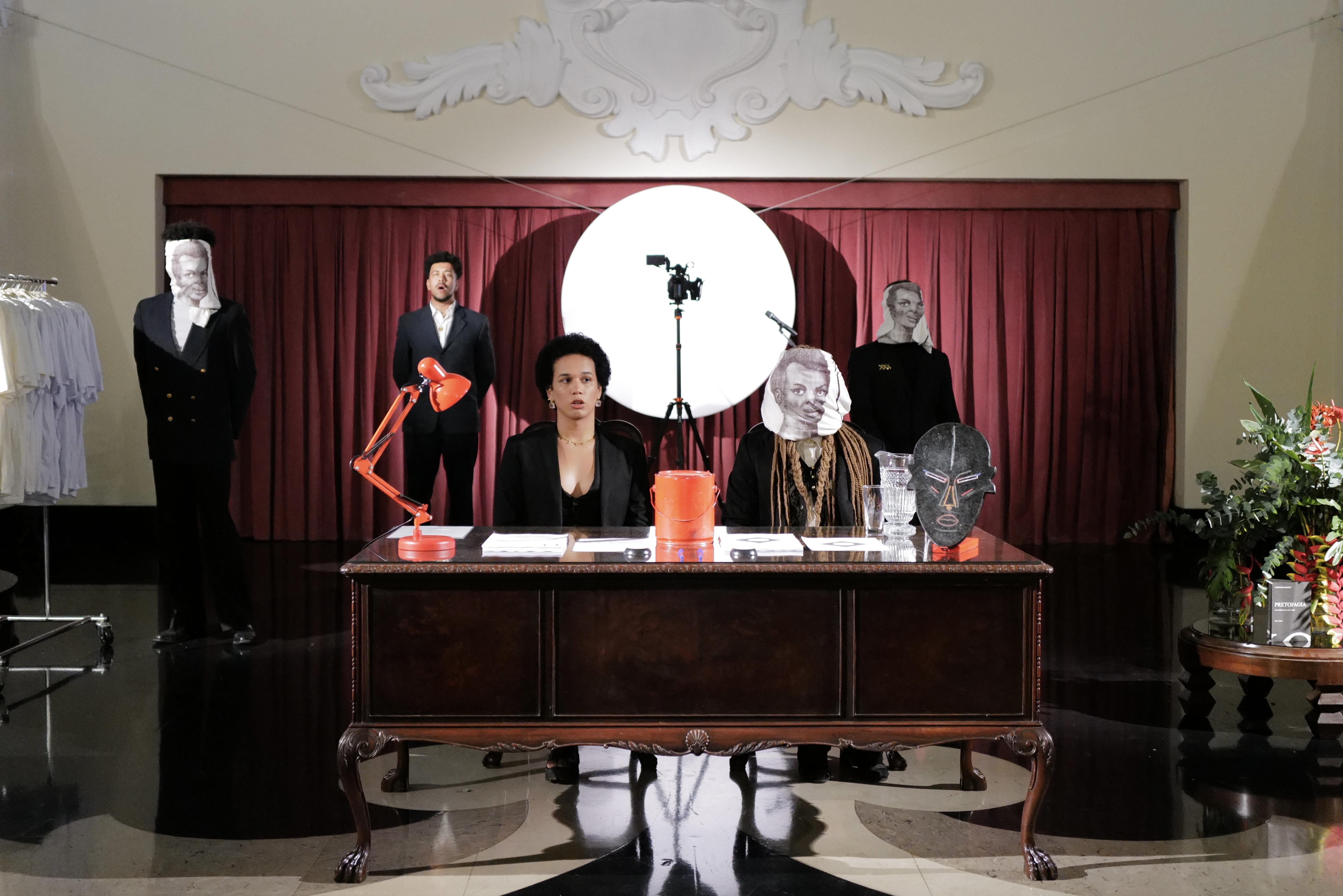 In an ornately furnished room with a red curtain in the background, there are five performers in black suits. Two sit behind a wooden desk that holds office supplies and an African mask. One of the performers at the desk wears a white cloth wrapped around their head with a face printed on the front. Behind them stand three other performers, two of whom wear similar clothes and one of whom is wearing the same cloth mask. Behind the performers in the center of the composition sits a circular reflector panel with a camera on a tripod positioned in front of it. To the left of the scene is a clothing rack filled with white t-shirts and to the right a wooden table with plants.