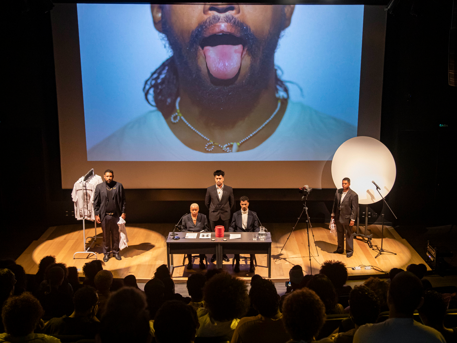 In a theater, five performers dressed in black suits stand on a stage. At the center, two people are seated behind a desk while another stands between them. Behind this scene, a large screen displays a close up image of a person wearing a white t-shirt and necklace with their tongue sticking out. To the left, a performer individual stands in front of a clothing rack of white t-shirts and to the right, a performer stands in front of a circular reflector panel. There are two bright spotlights shining down on the performers, adding an element of drama to the scene. The audience is visible in the foreground.