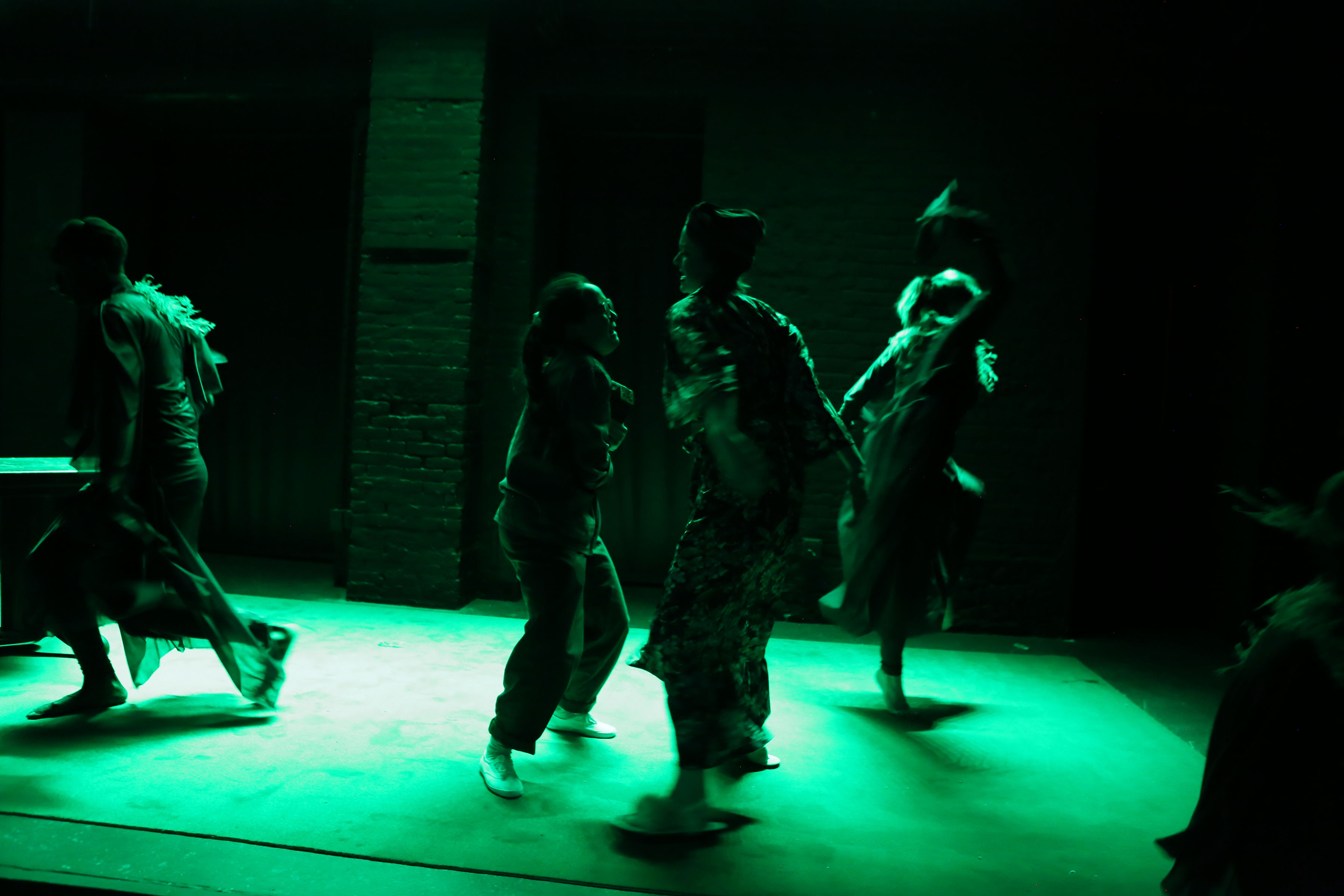 Four performers, Ashley Morton, Dorothea Gloria, Alice Lussiana Parente, and Shawn Herb Felton, are captured mid-movement on a stage, all shown in profile rather than facing the camera. Two stand near the center, turned toward one another, while one performer at downstage right balances on one foot as if stepping out of the frame, and another at upstage left is also about to exit. The stage is washed in green light, casting a vivid green hue across the scene, with a bright, light-colored floor—likely white marley—reflecting the green illumination.