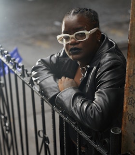 Portrait of Kearra Amaya Gopee. Gopee stands on a sidewalk, looking into the camera while resting their arms on a black iron gate. They are wearing a black leather jacket, sunglasses with a thick rectangular white frame, a silver chainmail necklace, a silver hoop earring, a silver curved barbell eyebrow ring, and black lipstick.